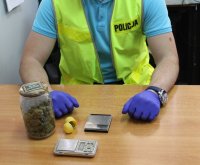 policjant siedzący przy biurku zabezpieczający znalezione w trakcie przeszukania przedmioty słoik, plastikowe opakowanie zawierające susz roślinny i wagi