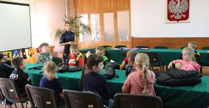 policjantka stoi w auli budynku komendy Policji w Wieluniu i prowadzi pogadankę wśród dzieci siedzących przy stole.