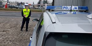 Policjantka stoi przy drodze i nadzoruje ruch , w ręku trzyma tarczę do zatrzymywania pojazdów, z boku częściowo widoczny radiowóz.