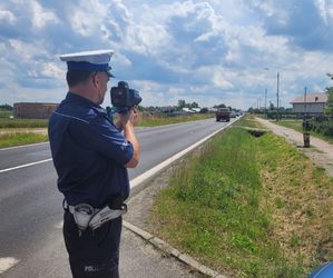Umundurowany policjant ruchu drogowego pełni służbę na drodze, dokonuje pomiaru ręcznym miernikiem prędkości.
