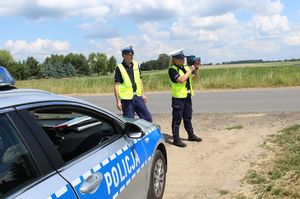 Dwóch umundurowanych policjantów ruchu drogowego pełni służbę na drodze. Jeden z nich dokonuje pomiaru ręcznym miernikiem prędkości. Z boku stoi radiowóz.