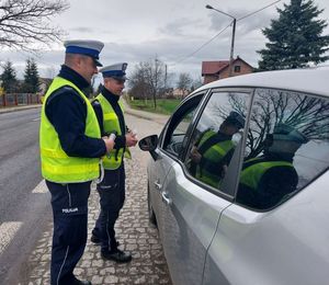 Policjanci prowadzą kontrolę drogową wręczają kierowcy zawieszkę zapachową.