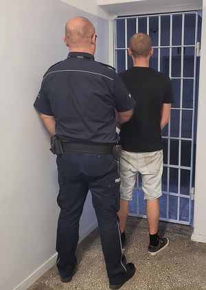 Policjant prowadzi zatrzymanego po korzytarzu w pomieszczeniu dla osób zatrzymanych.