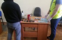 kolorowy obrazek przedstawiający dwie osoby stojące w pomieszczeniu przy biurku. Jeden z nich ma założoną kamizelkę z napisem policja. Na biurku leżą zabezpieczone przedmioty.