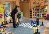Policjantka z przedszkolakami w trakcie pogadanki edukacyjno- profilaktycznej na temat bezpieczeństwa. Policjantka uczy dzieci  prawidłowego przechodzenia przez jezdnię.