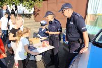 radiowóz stojący w tle oraz policjantka wraz z policjantem na Jesiennym Pikniku, która przeprowadza quiz wiedzy o bezpieczeństwie wśród uczestników. Całość imprezy odbywa się na na powietrzu.