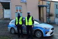 zdjęcie przedstawiające parking przed wieluńską komendą Policji i zaparkowany przed budynkiem radiowóz policyjny. Przed radiowozem stoją dwaj umundurowani policjanci drogówki.