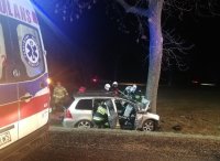 na zdjęciu widoczna częściowo karetka pogotowia oraz pojazd na poboczu drogi, który uderzył w drzewo. Strażacy udzielający pomocy poszkodowanemu.