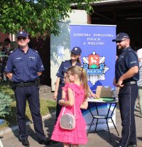 Policjanci w trakcie wydarzenia plenerowego &quot;Emdekolada&quot; na placu Powiatowego Młodzieżowego Domu Kultury i Sportu w Wieluniu. Wystawione stoisko profilaktyczne, wraz z banerem Komendy Powiatowej Policji w Wieluniu. Obok stoi zaparkowany radiowóz. Umundurowani policjanci przeprowadzają quiz dla dzieci dotyczący bezpieczeństwa i wręczają elementy odblaskowe.