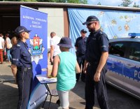 Policjanci w trakcie wydarzenia plenerowego &quot;Emdekolada&quot; na placu Powiatowego Młodzieżowego Domu Kultury i Sportu w Wieluniu. Wystawione stoisko profilaktyczne, wraz z banerem Komendy Powiatowej Policji w Wieluniu. Obok stoi zaparkowany radiowóz. Umundurowani policjanci przeprowadzają quiz dla dzieci dotyczący bezpieczeństwa i wręczają elementy odblaskowe.