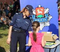 Policjantka w trakcie wydarzenia plenerowego &quot;Emdekolada&quot; na placu Powiatowego Młodzieżowego Domu Kultury i Sportu w Wieluniu. Wystawione stoisko profilaktyczne, wraz z banerem Komendy Powiatowej Policji w Wieluniu.  Wręcza element odblaskowy dziecku.