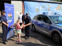 Policjanci w trakcie wydarzenia plenerowego &quot;Emdekolada&quot; na placu Powiatowego Młodzieżowego Domu Kultury i Sportu w Wieluniu. Wystawione stoisko profilaktyczne, wraz z banerem Komendy Powiatowej Policji w Wieluniu. Obok stoi zaparkowany radiowóz. Umundurowani policjanci przeprowadzają quiz dla dzieci dotyczący bezpieczeństwa i wręczają elementy odblaskowe.