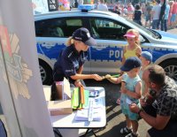 Policjanci w trakcie wydarzenia plenerowego &amp;quot;Emdekolada&amp;quot; na placu Powiatowego Młodzieżowego Domu Kultury i Sportu w Wieluniu. Wystawione stoisko profilaktyczne, wraz z banerem Komendy Powiatowej Policji w Wieluniu. Obok stoi zaparkowany radiowóz. Umundurowani policjanci przeprowadzają quiz dla dzieci dotyczący bezpieczeństwa i wręczają elementy odblaskowe.