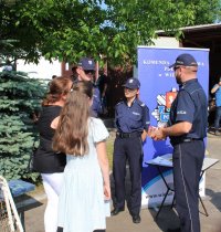 Policjanci w trakcie wydarzenia plenerowego &quot;Emdekolada&quot; na placu Powiatowego Młodzieżowego Domu Kultury i Sportu w Wieluniu. Wystawione stoisko profilaktyczne, wraz z banerem Komendy Powiatowej Policji w Wieluniu. Obok stoi zaparkowany radiowóz. Umundurowani policjanci przeprowadzają quiz dla dzieci dotyczący bezpieczeństwa.