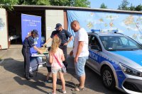 Policjanci w trakcie wydarzenia plenerowego &quot;Emdekolada&quot; na placu Powiatowego Młodzieżowego Domu Kultury i Sportu w Wieluniu. Wystawione stoisko profilaktyczne, wraz z banerem Komendy Powiatowej Policji w Wieluniu. Obok stoi zaparkowany radiowóz. Umundurowani policjanci przeprowadzają quiz dla dzieci dotyczący bezpieczeństwa i wręczają elementy odblaskowe.