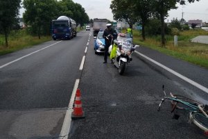 Zdjęcie przedstawia policjantów pracujących na miejscu wypadku. Na zdjęciu droga gdzie doszło do zdarzenia, rower, samochód ciężarowy,  policjanci, policyjny motocykl, wóz strażacki