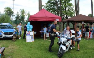 motocykl policyjny na którym siedzą dzieci obok stoi policjant. W tle stoisko profilaktyczne i radiowóz