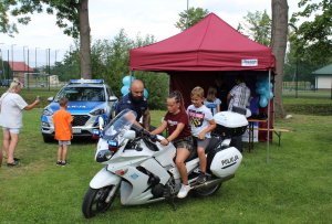 motocykl policyjny na którym siedzą dzieci obok stoi policjant. W tle stoisko profilaktyczne i radiowóz