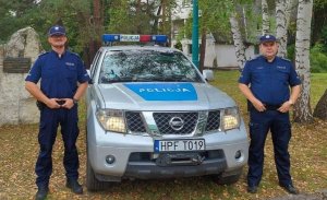 dwaj umundurowani policjanci stoją po bokach radiowozu