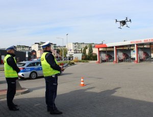 startujący dron, obok dwóch policjantów jeden z nich jest operatorem drona, w tle radiowóz