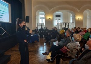 policjantka stoi , mówi do mikrofonu,  na krzesłach siedzą goście oraz seniorzy w sali Wieluńskiego Domu Kultury
