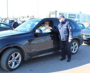 policjant na parkingu wręcza kierowcy siedzącemu w aucie elementy odblaskowe i zawieszki zapachowe przechodniom