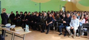 w sali gimnastycznej w szkole na krzesłach siedzi młodzież oraz w pierwszym rzędzie zaproszeni goście, policjantka stoi na przeciwko informując o celu debaty.