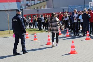 przed halą sportową na parkingu ustawiony jest z pachołków tor przeszkód, który pokonywany jest przez uczennicę obok stoi policjant w tle są inni uczniowie.