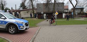 miasteczko ruchu drogowego, z boku stoi radiowóz. Po ulicach miasteczka jedzie uczeń na rowerze, policjant stoi w tle ocenia jazdę. W tle widoczni uczestnicy turnieju.