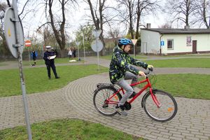 miasteczko ruchu drogowego, po jego  ulicach  jedzie uczeń na rowerze pokonując przeszkody, policjant stoi w tle ocenia jazdę.