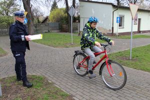 miasteczko ruchu drogowego, po jego ulicach  jedzie uczestnik turnieju na rowerze, policjantka stoi w tle ocenia jazdę.