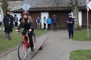 miasteczko ruchu drogowego, po jego ulicach  jedzie uczestnik turnieju na rowerze, pokonując równoważnię  policjantka stoi w tle ocenia jazdę