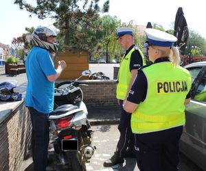 policjanci stoją na parkingu gdzie zaparkowany jest motocykl obok stoi motocyklista prowadzą z nim rozmowę na temat bezpieczeństwa.