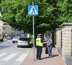 policjantka stoi na chodniku rozmawia z pieszą, w tle widoczna droga, przejście dla pieszych oraz znak informujący o przejściu.