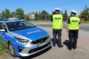 policjanci stoją przy drodze w tle radiowóz. Jeden z policjantów mierzy prędość