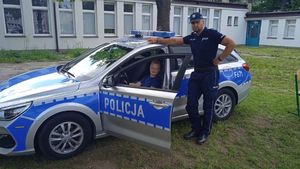 policjant stoi przy radiowozie, w którym otwarte są drzwi. Na siedzeniu kierowcy siedzi dziecko.