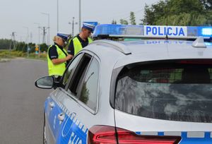 Umundurowany policjant i policjantka stoją przy drodze na tle radiowozu.