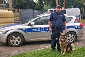 Pies służbowy wraz z przewodnikiem pozują do zdjęcia na tle radiwozu.