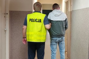 Policjant w kamizelce z napisem Policja prowadzi zatrzymanego po korytarzu budynku komendy.