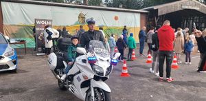 Plac na zewnątrz terenie domu kultury, motocykl policyjny na którym siedzi chłopiec, z boku stoi umundurowany policjant ruchu drogowego, w tle stoisko profilaktyczne Policji oraz uczestnicy imprezy.