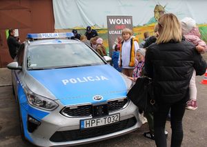 Plac na  terenie domu kultury, w  tle stoisko profilaktyczne Policji i radiowóz policyjny wokół stoją uczestnicy imprezy.