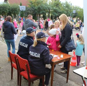 Plac na zewnątrz terenie domu kultury,  przy stoliku siedzą policjanci wykonują dzieciom pamiątkowe odbitki linii papilarnych.