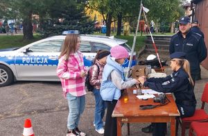 Plac na zewnątrz terenie domu kultury,  przy stoliku siedzi policjantka i wykonuje dzieciom pamiątkowe odbitki linii papilarnych.