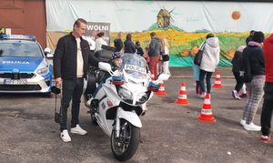 Plac na zewnątrz terenie domu kultury, motocykl policyjny na którym siedzi chłopiec,  w tle stoisko profilaktyczne Policji oraz uczestnicy imprezy.