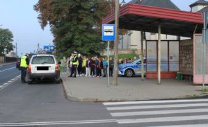 Droga, policjant zatrzymał pojazd do kontroli, z boku na chodniku stoją dzieci, które uczestniczą w akcji Jabłko i cytryna.