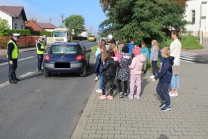 droga, policjant zatrzymał pojazd do kontroli, z boku na chodniku stoją dzieci, które uczestniczą w akcji Jabłko i cytryna