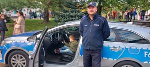 policjant stoi przy radiowozie, w którym na miejscu kierowcy siedzi chłopiec, w tle uczestnicy pikniku