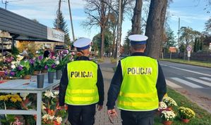 Policjanci idą chodnikiem wzdłuż drogi przy cmentarzu.