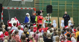 Policjanci wspólnie ze Świętym Mikołajem, Śnieżynką,efem i Myszką Miki stoją w sali gimnastycznej, jeden z policjantów mówi przez mikrofon, dzieci siedzą na przeciwko.