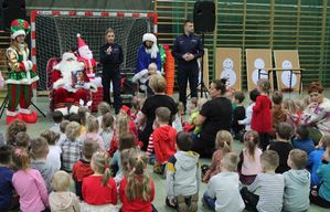 Policjanci wspólnie ze Świętym Mikołajem, Śnieżynką,efem  stoją w sali gimnastycznej,  policjantka mówi przez mikrofon, dzieci siedzą na przeciwko.